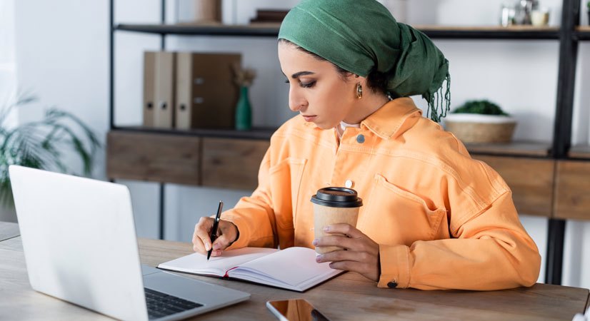 Woman wearing headscarf writing in book in front of a computer